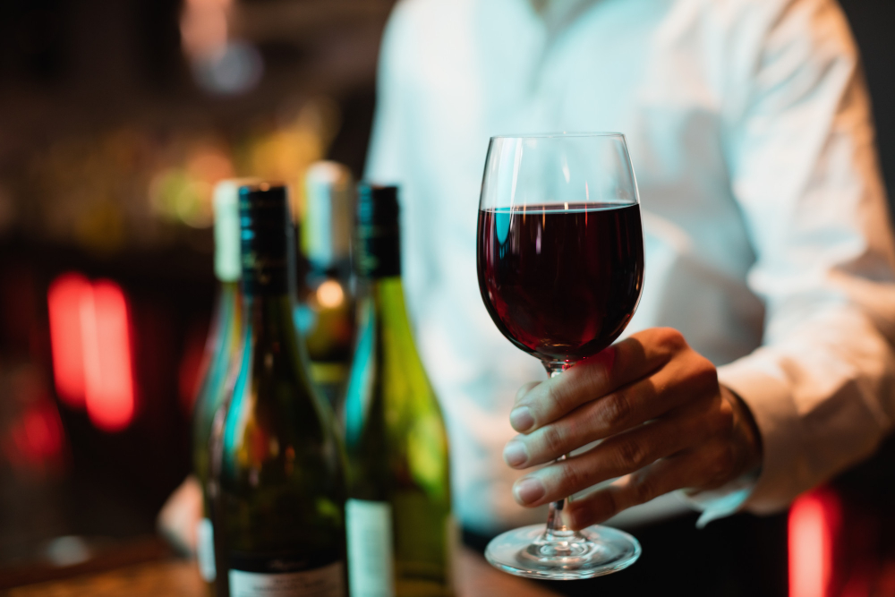Garçom oferecendo uma taça de vinho tinto com três garrafas colocadas no balcão e ambiente do retaurante desfocado ao fundo