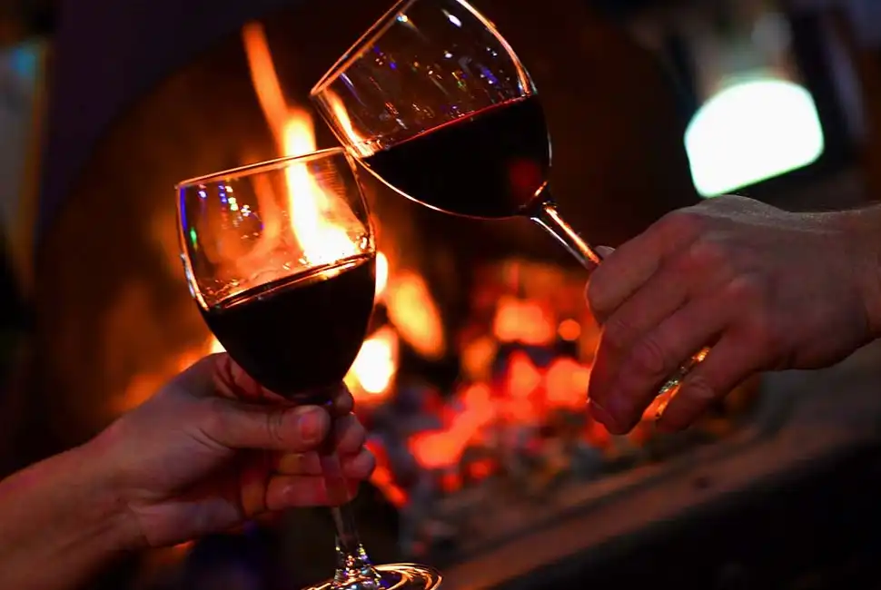 Foto em close de duas mãos brindando com duas taças de vinho com uma lareira ao fundo